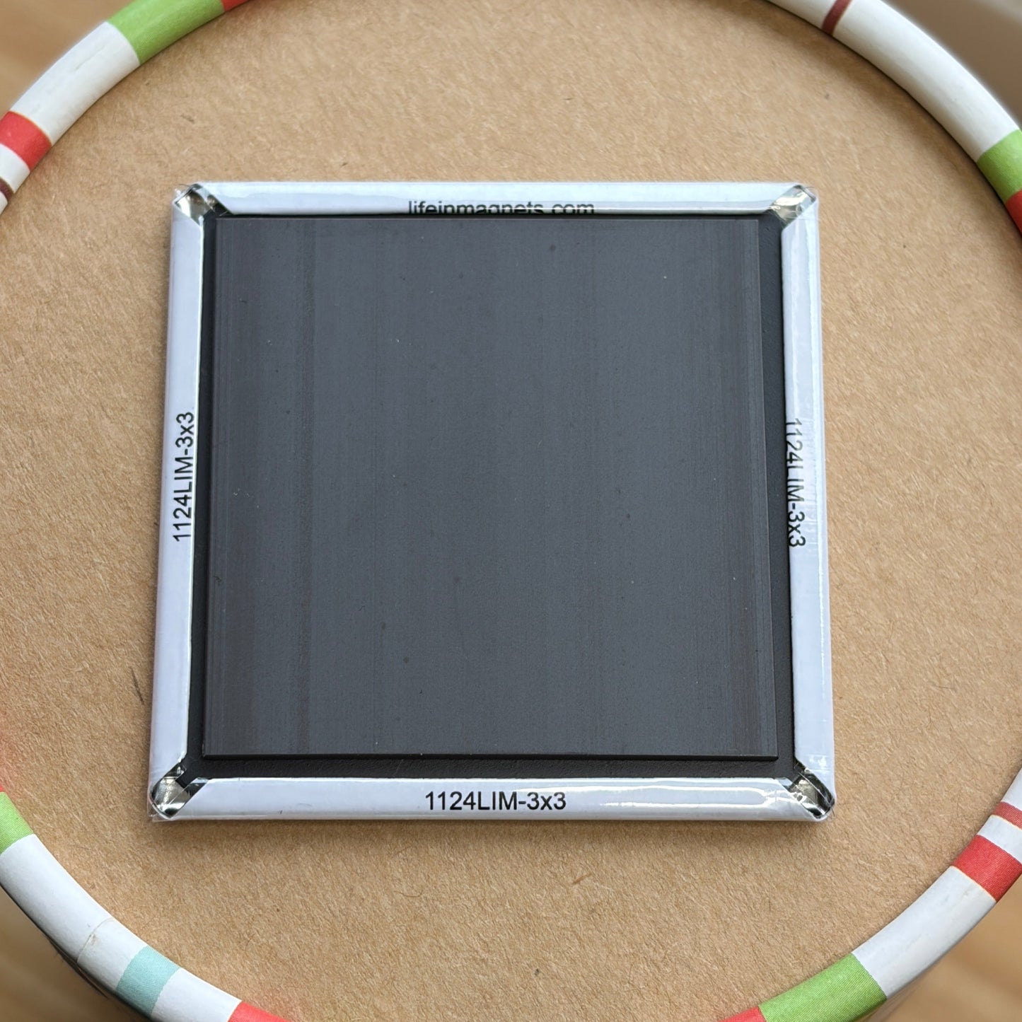 A square panel with a silver frame and dark gray/black surface, placed in the center of a circular ring made of white, red, green, and blue segments, on a brown cardboard surface.