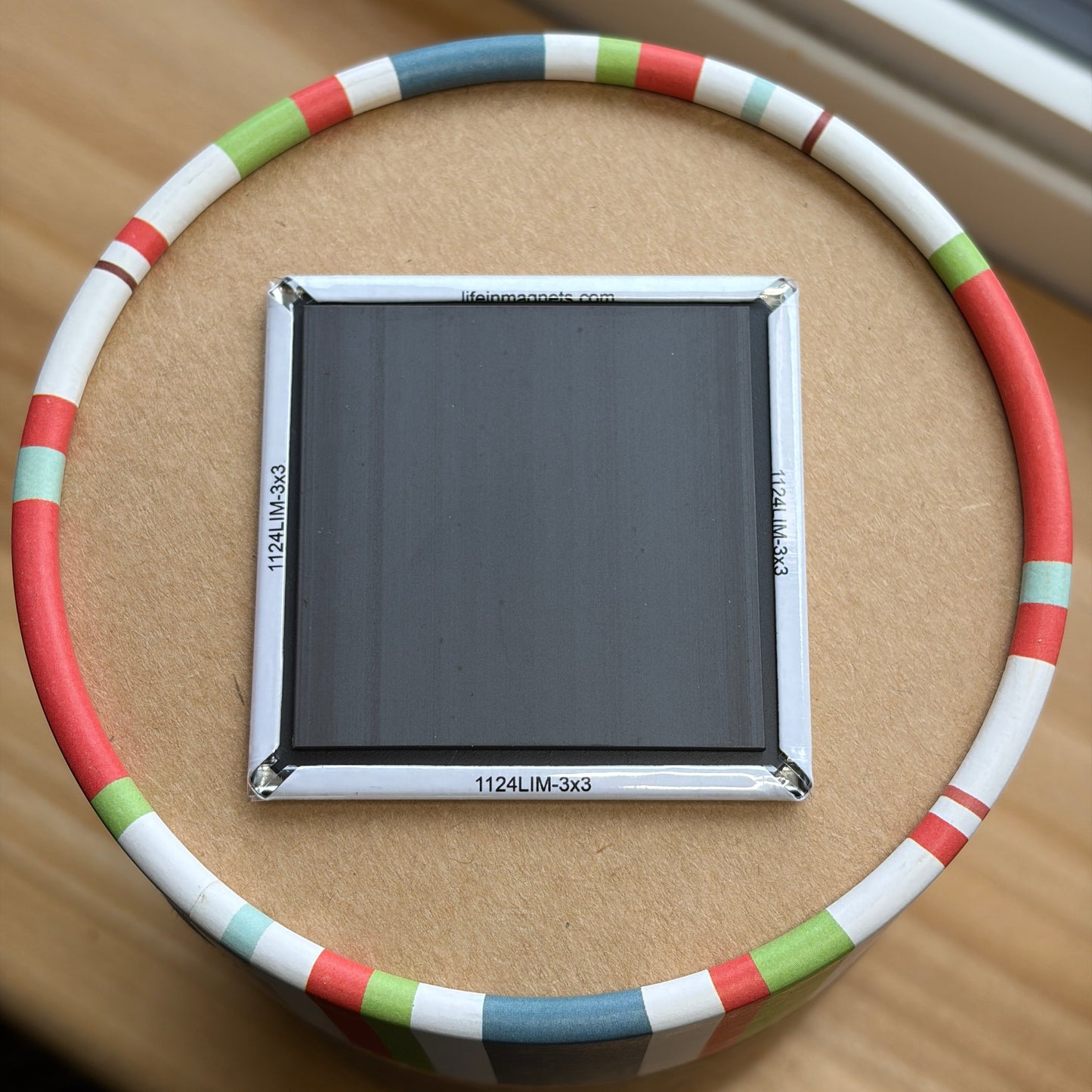 A square panel with a silver frame and dark gray/black surface, placed in the center of a circular ring made of white, red, green, and blue segments, on a brown cardboard surface.
