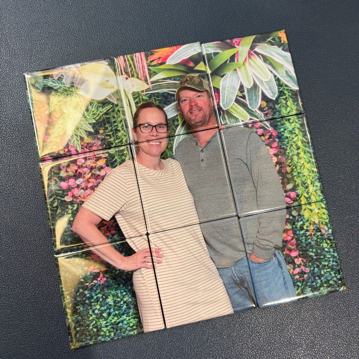 A 9-piece custom photo mosaic tile set arranged in a 3x3 grid, featuring a printed image of a smiling couple in front of a lush green garden wall, displayed on a dark textured surface.