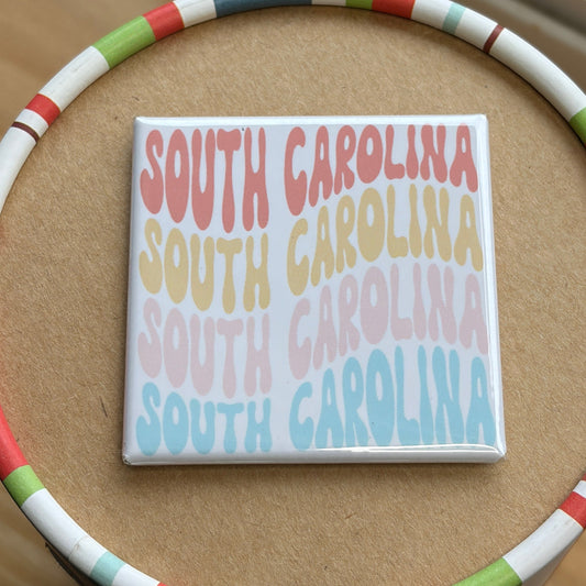 A square coaster featuring the text 'SOUTH CAROLINA' repeated in pastel red, orange, yellow, pink, and blue, displayed on top of a round cardboard tray with a colorful striped rim.